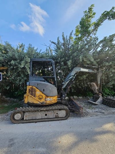 A yellow and gray mini excavator is parked on a gravel and asphalt surface in front of a dense green hedge under a blue sky with scattered clouds. The excavator is positioned at an angle to the right, with its tracks on the ground. The cab of the excavator is open at the top, and its boom is extended. A bucket is attached to the boom, and another bucket is visible on the ground to the right of the excavator. A portion of a tire is also on the ground to the right. The overall scene suggests an outdoor setting, possibly a construction site or a storage area, in Islamorada, United States of America.