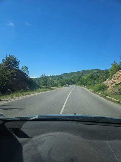 The image shows a road in Liaskovo, Bulgaria, as seen from inside a vehicle. The road is asphalt and has a white line marking the center. To the left of the road is a steep, rocky hillside covered with green vegetation. The sky is clear blue. The interior of the vehicle, including part of the dashboard, is visible on the right side of the frame.