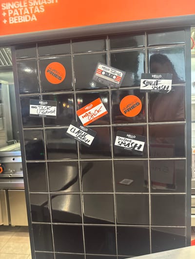 The image shows a wall covered in black square tiles. Several stickers are affixed to the tiles.
At the top of the image, an orange sign with white text reads "SINGLE SMASH + PATATAS + BEBIDA".
On the tiles, from top left to bottom right, the stickers are:
- A circular orange sticker with black text that reads "UNLIMITED FRIES" and a cartoon face.
- A rectangular sticker designed to look like a cassette tape with "HELLO MY NAME IS BACON SMASH" printed on it.
- A rectangular white sticker with "HELLO MY NAME IS SINGLE SMASH" and "@BASICSBYGOIKO" printed at the bottom.
- A rectangular white sticker with "HELLO MY NAME IS BIG JACK SMASH" and "@BASICSBYGOIKO" printed at the bottom.
- A rectangular orange sticker with white text that reads "HELLO MY NAME IS HOT CHICK" and "@BASICSBYGOIKO" printed at the bottom.
- A rectangular white sticker with "HELLO MY NAME IS CLASSIC SMASH" and "@BASICSBYGOIKO" printed at the bottom.
- A rectangular white sticker with "HELLO MY NAME IS BASIC ONION SMASH" and "@BASICSBYGOIKO" printed at the bottom.
The background includes reflections on the tiles and a hint of kitchen equipment to the left. The setting appears to be a food establishment. This image was taken in Arroyomolinos, Spain.