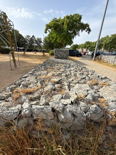 The image depicts an outdoor scene in a park-like area with a playground. In the foreground, a gabion wall constructed from large rocks and wire mesh runs diagonally across the frame, with dry grass and weeds growing around it. Beyond the gabion wall, a sandy playground area is visible, featuring a jungle gym on the left and a triangular swing set structure in the center. A child is seen on one of the swings. In the background, a paved walkway runs alongside another gabion wall, leading to a parking lot filled with cars. Several trees with dense green foliage are present throughout the scene, and the sky is partly cloudy. The location is identified as the Parc de l'Amphitheatre in Mataró, Spain.