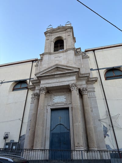 Here is a detailed description of the image:

The image captures an angled view of a building facade in Acireale, Italy. The architectural style suggests a historical or religious structure, likely a church or similar edifice. 

The facade is characterized by elaborate stonework, including decorative columns with intricate capitals, a triangular pediment above the entrance, and a square bell tower with an arched opening. A blue door is prominent and features an embossed cross. A small plaque mounted on the stone facade contains Latin text. 

To the right of the entrance is a metal fence. A lit star hanging from the roof is visible.

The sky is visible above the building, indicating daytime. A black power cable is prominent on the left side. The building has windows with rounded tops.
