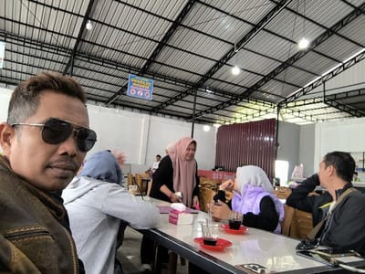 This media file depicts a group of people gathered at a table in what appears to be an outdoor or semi-outdoor dining area, likely a cafe or restaurant in Panteraya, Indonesia. The setting features a corrugated metal roof supported by a black metal framework, with industrial-style lighting fixtures. The overall atmosphere suggests a casual daytime gathering.

In the foreground, a man wearing sunglasses and a brown leather jacket is looking towards the camera, taking up a significant portion of the frame. He has a mustache and short, styled hair. Behind him, several individuals are seated around a table. One woman in a pink hijab is smiling and gesturing while holding a smartphone and a pink box of tissues. Another person wearing a light blue hijab and a grey hoodie is also seated at the table. Across from them, a woman in a purple vest and hijab is holding a drink, and a man in a black jacket is looking away, resting his head on his hand.

On the table are several items, including glasses with dark liquid (likely coffee or tea), red saucers, straws, a pink tissue box, and what looks like a smartphone. The table itself is white and appears to be a laminate surface.

In the background, there are other tables and chairs, suggesting a public dining space. A maroon-colored wall is visible on the right. There is also a sign hanging from the ceiling with Indonesian text, partially obscured but appearing to be a warning or informational sign with symbols. One visible sign has text that reads "DILARANG MEROKOK" which translates to "NO SMOKING." The lighting suggests it is daytime. The mood appears relaxed and social.