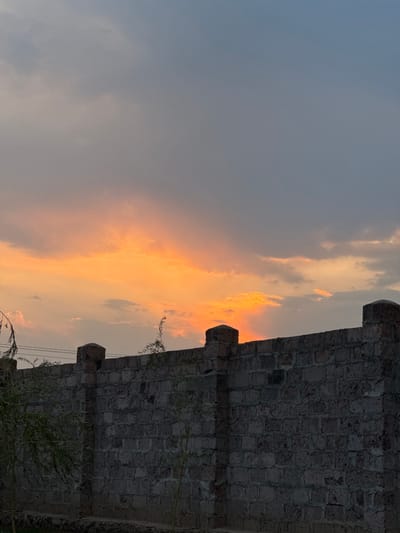The scene is located in Yeghvard, Armenia. The image captures a landscape at dusk or dawn. A substantial dark grey wall constructed of rectangular stone blocks, approximately two meters in height, extends across the lower portion of the frame. This wall features several uniformly spaced structural pillars topped with rounded caps. Sparse green foliage, appearing to be branches from a tree or large bush, is visible on the left side, extending into the frame and slightly over the wall's top. The upper half of the image is dominated by the sky, displaying a vivid gradient of color. The horizon area, positioned just above the wall, is illuminated with intense orange and yellow hues, indicative of a setting or rising sun. This coloration transitions upward into layers of grey and muted blue clouds, which cover the remaining sky. Prominent rays of bright orange light are visible ascending from behind the wall near the right side, piercing through the lower cloud cover. No people, animals, or discernible actions are present in the frame.