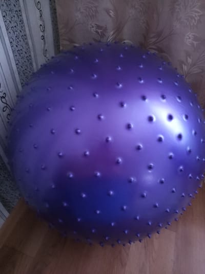 This media file features a large, purple, textured exercise ball as the primary object. The ball is covered in numerous small, raised bumps, giving it a spiky appearance. It is positioned on a wooden floor, and behind it, a patterned wallpaper and a vertical striped surface are visible, suggesting an indoor setting. The lighting appears to be artificial, casting highlights on the smooth sections of the ball and creating subtle shadows. There are no people or discernible activity taking place; the focus is solely on the exercise ball and its immediate surroundings. The scene is static and lacks any indicators of time of day, weather conditions, or specific emotional tone. No visible text is present in the image. The location context provided is Älimbet, Kazakhstan.