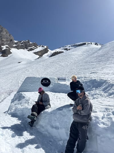 The image shows a snow-covered mountainous landscape in La Giettaz, France, with a bright blue sky above. Three people are present, appearing to be tourists or recreational skiers/snowboarders. One individual, wearing a grey hoodie and dark pants, is holding a phone and seems to be taking a picture. Another person is seen with blonde hair and white goggles, near a sign that reads "La Clusaz" (likely a sign for the ski resort or area). The third person has a pink hat and winter clothing. The ground is covered in snow, with visible tracks indicating previous ski or snowboard activity. A circular sign bearing the "Oakley" logo is also visible on the snow. The overall scene suggests a bright, sunny day at a ski resort in the French Alps.