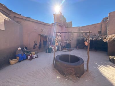 The scene depicts a meticulously constructed outdoor film set located within one of Ouarzazate, Morocco's film studios, likely Atlas Film Studios or CLA Studios. The environment is a traditional-style village courtyard, characterized by unpainted, reddish-brown earthen buildings with flat roofs, some featuring exposed thatch. The ground is compacted dirt. Bright, overhead sunlight illuminates the scene, casting distinct shadows and creating lens flare in the upper right quadrant, under a clear blue sky.
In the foreground, slightly to the right of center, a dark, oval-shaped well structure is prominent, equipped with a rustic wooden framework from which a rope and bucket descend. A terracotta pot hangs from one of the well's wooden supports.
On the left side of the courtyard, two figures, appearing to be static mannequins, are seated against a building wall. The figure on the left is draped in a blue garment and a grey shawl with a white head covering. The figure on the right wears a dark head covering and a green outer robe over darker clothing. In front of them, a low display stand holds several small, framed items. A woven straw basket rests on the ground beside them.
Further into the background, along the perimeter walls, are additional figures and structures resembling market stalls or workshops. These stalls feature thatched overhangs and display various items, including textiles and pottery. The overall scene suggests a historical or cultural tableau, designed for cinematic production.