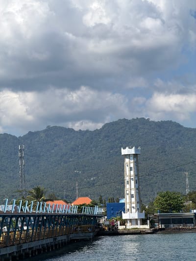 The image depicts a waterfront scene in Pemenang Timur, Indonesia, dominated by a lush, green mountain range in the background under a partly cloudy sky. In the foreground, a body of water reflects the sky and surrounding structures. A long, elevated pedestrian walkway with blue railings and yellow supports extends from the left side of the frame into the distance. To the right of the walkway, a tall, white, rectangular tower with several windows stands near the water's edge. Adjacent to the tower are several buildings with orange and blue roofs, and a dense cluster of trees. Further to the right, a building with a dark roof and signage reading "WARUNG MAKAN" and "KOKCHENG" is visible, situated on a rocky shoreline. A tall cell tower is also present in the middle ground, to the left of the mountain. The overall impression is that of a coastal area with active infrastructure and natural beauty.
