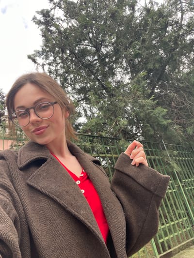 A female subject is depicted in an outdoor self-portrait in Cava de' Tirreni, Italy, under an overcast sky. She occupies the lower half of the frame, facing the camera with a subtle smile. She wears round, dark-framed eyeglasses, a dark brown herringbone-patterned overcoat with wide lapels, and a red v-neck top with white buttons. Her right hand holds the cuff of her left sleeve. Behind her, a large evergreen tree with dense, dark green foliage and visible branches rises against the sky. A dark green metal fence is visible in the background, extending from the center to the right, and a light pink building wall is partially visible on the far left. The precise location within Cava de' Tirreni cannot be determined from the visual evidence.