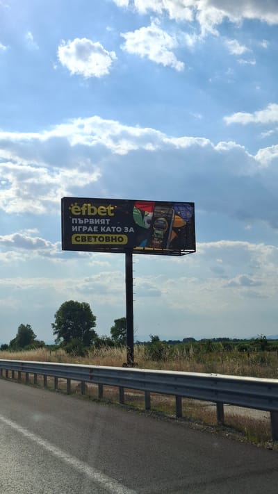 The image shows a billboard with the Efbet logo and text in Bulgarian. The billboard is located in Malo Konare, Bulgaria, alongside a highway. The billboard advertises Efbet, a betting company. The text on the billboard reads "първият играе като за световно", which translates to "the first plays like for the world". The billboard features an image of a mobile phone displaying betting options for different countries. The background consists of a blue sky with white clouds, trees, and fields. A guardrail is visible in the foreground, separating the highway from the roadside. The lighting suggests it is daytime, possibly late afternoon due to the direction of shadows. No people are visible in the image.