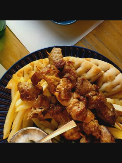 The image shows a close-up of a dish of souvlaki, fries, and pita bread served on a dark blue plate. The souvlaki appears to be made of grilled pork skewers. The fries are golden brown and appear to be thin-cut. The pita bread is flat and slightly puffed. There is a small bowl of white sauce, possibly tzatziki, visible at the bottom left of the plate with a spoon in it. The food is arranged on a wooden table, and a white surface is visible in the background. The lighting suggests it might be daytime. The scene is captured in Bielawska Wieś, Poland.