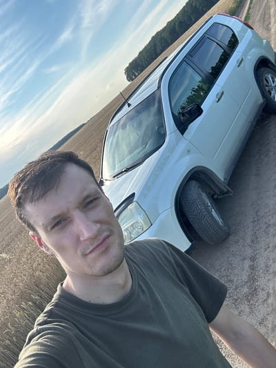 A man in a green t-shirt is taking a selfie next to a white Nissan X-Trail SUV parked on a dirt road. The car is positioned at an angle to the right, with its front left wheel slightly turned. The man's face is partially visible in the foreground, looking directly at the camera. Behind the car, a field of ripe wheat stretches towards a treeline, and beyond that, a clear blue sky with wispy clouds. The image appears to have been taken during daylight, likely in the late afternoon due to the quality of light. The location is the village of Slobidka-Zalisetska, Lviv Oblast, Ukraine.