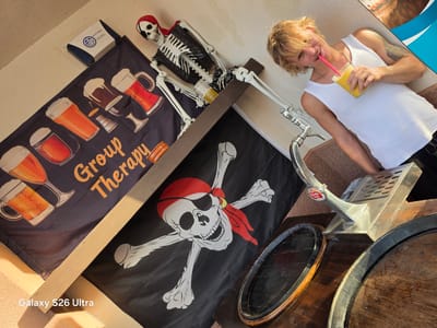 The image shows a person with blond hair, wearing a white tank top and dark pants, holding a yellow drink with a pink straw and smiling. They are standing next to a skeleton dressed as a pirate, complete with a red bandana. Behind them are two flags: one depicting various beers and the text "Group Therapy," and another with a skull and crossbones. The scene appears to be outdoors, with sunlight casting shadows, suggesting it is daytime. Wooden barrels and a beer tap are visible, hinting at a bar or pub setting. The location is identified as Llucmajor, Spain. The overall mood seems jovial and celebratory.