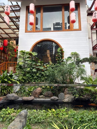 A partial exterior view depicts a commercial establishment within An Hải Ward, Vietnam. The central structure features a two-story facade primarily clad in white horizontal siding. The upper level includes four rectangular windows with brown wooden frames, reflecting an overcast sky and the dark silhouette of an adjacent multi-story building. Below these, a large circular window with a prominent brown wooden frame is centered on the lower facade, reflecting interior lighting and structural elements.

Six cylindrical paper lanterns are suspended: three red lanterns hang from a translucent overhead structure on the left, and three (two light pink, one red) are suspended from the white facade above the rectangular windows. Each visible lantern displays Japanese characters, specifically "寿司" (sushi). To the immediate left, a dark brown wooden railing and a section of an upward-sloping staircase are visible, adorned with additional small red lanterns and red decorative ribbons. On the far right, a section of an adjacent structure with exposed reddish metal support beams and a rough concrete/stone wall is partially visible.

The foreground features a landscaped garden area. A dark-water pond occupies the lower-center, containing several golden-orange koi fish visible near the right edge. The pond is bordered by various natural rocks of differing sizes and shades of grey and brown. A low barrier, constructed from a metal frame and transparent panels, horizontally traverses the midground, separating the pond from deeper garden beds. These beds contain dense vegetation, including large-leafed tropical plants, tall slender-leaved plants resembling papyrus, and various ground cover plants. No individuals are present in the scene. The lighting indicates overcast or soft daylight conditions.