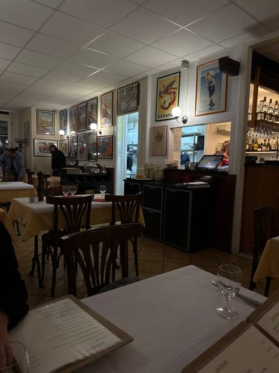 The image depicts an interior view of a restaurant, possibly located in Chêne-Bourg, Switzerland given the query context. The restaurant has a bar area visible towards the back, with bottles and glassware displayed. A serving hatch connects the bar/kitchen area to the dining space. Walls are adorned with a series of framed posters or prints, which vary in size and theme, running along the length of the dining area. Diners or restaurant staff can be seen in the background, and at least one person is at a table. The foreground is dominated by a table setting, covered with a white tablecloth, a menu, and glasses. Wooden chairs are placed around the tables. The lighting appears to be a mix of ambient light and focused lighting on the artwork and tables. The overall atmosphere is casual and intimate.