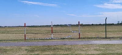 The image captures a wide, open outdoor landscape under a bright, clear blue sky with scattered white clouds. In the foreground, a dark paved path or road cuts across the bottom, separating a narrow strip of dry grass from a larger, expansive grassy field that extends into the horizon.

In the midground, a series of three distinct posts or poles create a barrier-like structure. On the left, a prominent red and white striped pole stands upright. To its right, another white pole, also with visible red stripes, supports a loosely strung, light-colored net or barrier, which appears somewhat tattered and saggy in the middle. This net stretches between the two striped poles. Further to the right, a plain dark green post with a slight curve at its top completes the line of markers.

The vast grassy field behind these structures appears dry and sun-baked, with varying shades of green and brown. In the far distance, a faint line of vegetation, possibly trees or distant structures, can be seen along the horizon. The scene is brightly lit, indicating daytime, likely mid-day, with sunny and fair weather. There are no people visible, and no specific activity is taking place. The setting, particularly with the type of marker poles and extensive open field, suggests a large open area such as an airfield or a vast park, consistent with the San Fernando, Argentina, location context. No text is visible in the image.