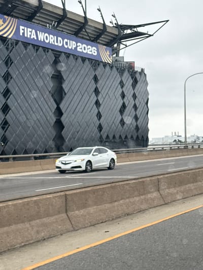 This is a vertical, eye-level, medium shot capturing a modern building with a large banner and a white car driving on a road next to it. The building's facade is covered in a diamond-patterned, dark material, with some scaffolding visible at the top. A prominent blue banner with gold lettering and stars hangs on the left side of the building, displaying "FIFA WORLD CUP 2026". The car is a sleek, white sedan in motion, its headlights illuminated. The scene appears to be outdoors, with a cloudy sky suggesting an overcast day, possibly morning or afternoon. The road is asphalt with white lane markings, separated from the building by a concrete barrier. The setting suggests an urban environment, likely a highway or major road adjacent to a significant structure or stadium. No people are clearly visible, but the presence of the car and the banner implies ongoing activity related to the upcoming FIFA World Cup. The overall impression is one of anticipation and modernity.