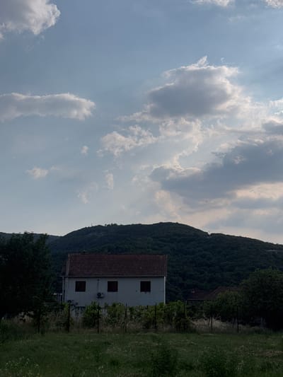 This image depicts a serene landscape scene in Radanovići, Montenegro. The primary subjects are a two-story house with a red tiled roof and a steep, densely forested mountain that forms the backdrop. The house is situated in the foreground on the right side of the frame, appearing to be unoccupied. Its facade is light-colored, with visible dark-framed windows and an air conditioning unit.
The scene is set outdoors, with a grassy area in front of the house and a path or driveway leading towards it, partially obscured by dark green trees. The mountain, covered in lush vegetation, dominates the upper right portion of the image. The sky above is partly cloudy, with streaks of sunlight breaking through, suggesting it might be late afternoon or early evening. The overall atmosphere is peaceful and tranquil, with no activity taking place.
Notable details include the clear division between the cultivated area around the house and the wilder mountain, hinting at a rural or semi-rural setting. The lighting suggests a pleasant, perhaps warm day. There is no visible text in the image. The orientation of the image is portrait.