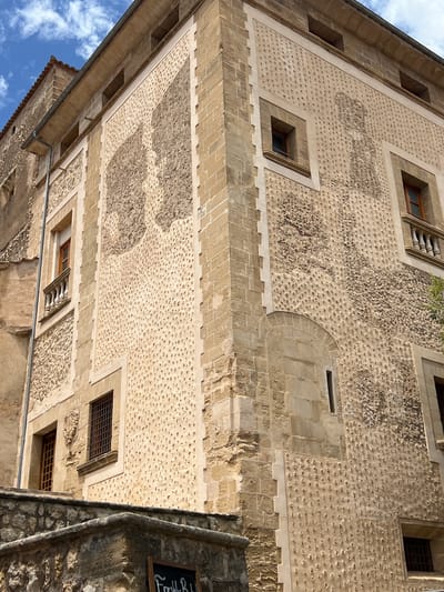 The image shows the exterior of a building in Pollença, Spain, with a distinctive textured facade. The building is constructed with light-colored stone blocks and features two prominent sections of wall decorated with numerous small, indentations or protrusions. These textured areas are arranged in irregular patterns, creating a rough, almost organic surface.

There are several windows visible, some with wooden frames and metal grilles, while others have small balconies with balustrades. The building's architecture suggests historical significance, with visible stone detailing around the windows and corners. Part of an adjacent building, also made of stone, is visible on the left, with a terracotta-tiled roof.

In the foreground, there is a stone wall or parapet, and a dark sign with white lettering is partially visible, suggesting a commercial establishment. The sky above is partly cloudy with a blue sky. The overall impression is of an old, well-preserved building in a Spanish town.