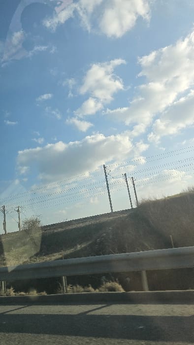 The image depicts a daytime outdoor scene in Stelnica, Romania. The upper two-thirds of the frame show a clear blue sky interspersed with scattered white cumulus clouds, illuminated by sunlight. In the foreground, a dark asphalt road surface is partially visible at the bottom. A silver-colored metal guardrail, composed of horizontal beams supported by vertical posts, runs horizontally across the lower mid-section of the image. Behind the guardrail, a sloped embankment rises, covered in dry, light brown and green grasses and sparse vegetation. Three dark-colored utility poles, each carrying numerous thin horizontal wires, are positioned along or just above the crest of the embankment, silhouetted against the sky. A small, leafless tree or shrub is visible on the embankment to the left. No individuals or active subjects are present in the scene.