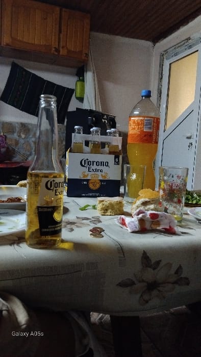 The scene depicts an indoor setting, specifically a dining or gathering area in Oreshene, Bulgaria. A light-colored tablecloth with a recurring pattern of light brown floral designs covers a table, which is the primary focus.
On the table, a clear glass bottle of "Corona Extra" beer stands upright, appearing partially consumed. To its right, a cardboard six-pack carrier for "Corona Extra" beers is present, containing several bottles. A large, clear plastic bottle with a blue cap holds an orange-colored beverage, likely soda or juice, with a partially visible label containing Cyrillic script. Two drinking glasses are positioned near the orange beverage bottle; one is clear, and the other features a colorful floral pattern.
Food items visible on the tablecloth include two pieces of a light-colored, crumbly cake or bread, one appearing as a cut slice and the other as a broken fragment. A crumpled white cloth or napkin with red markings lies between the cake and the floral glass. Two metallic coins, consistent with Bulgarian Leva, are placed on the tablecloth. A few green leaves are scattered on the surface. A white plate with remnants of food is partially visible on the far left.
The background features a textured wall on the left, possibly stone or stone-effect, above which two light-colored wooden wall cabinets are mounted. A dark, patterned textile hangs on the wall below the cabinets. To the right, a white PVC window or door frame is visible, leading to an illuminated area beyond. Above the beer carrier, a dark electronic device, possibly a speaker or radio, is situated on a shelf or surface. The ceiling is paneled with dark wooden slats. No individuals are visible in the image. The general lighting suggests an indoor setting, possibly during the evening.
