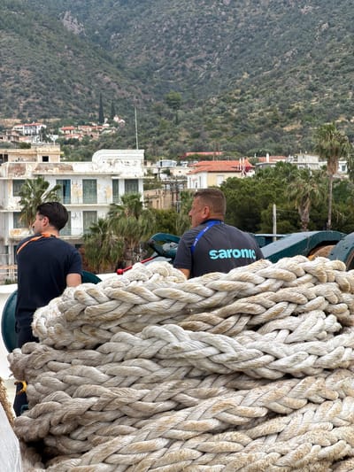 Two men are working with a large coil of rope on a ship docked in the Municipal Unit of Methana, Greece. The man closer to the camera wears a dark blue shirt with an orange strap over his shoulder. The man further away wears a dark gray shirt with the word "saronic" in blue. Buildings and trees are visible in the background, along with a hillside covered in green vegetation.