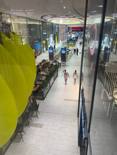 The image shows a brightly lit, modern shopping mall with multiple levels and a high, open ceiling. The flooring is tiled in a light color, and there are numerous shops and businesses visible along the walkways. A large, vibrant yellow sculptural element dominates the left side of the foreground, reflecting the mall's interior.
In the middle ground, two young women are walking away from the camera down a wide walkway. The woman on the left is wearing a pink t-shirt and pink shorts, and the woman on the right is wearing a white tank top and a white skirt. They are walking past a row of tables and chairs set up outside a restaurant or cafe area.
Further back, other shoppers are visible walking and interacting within the mall. Signs for businesses like "MediaMarkt" are prominent. The mall features large glass storefronts, modern lighting fixtures, and decorative greenery along some of the shopfronts. The overall impression is a busy, contemporary retail environment.
The image is identified as being taken in Aachen, Germany.