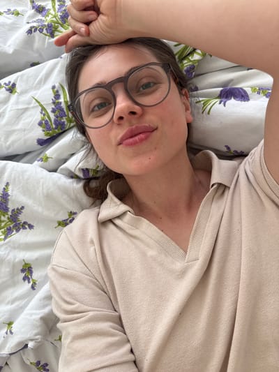 A person lies on their back on a bed with white floral-patterned sheets. The person wears glasses and a light-colored shirt. The location cannot be determined based solely on the image.