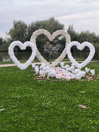 The image depicts an outdoor marriage proposal setup. Three large heart-shaped floral arrangements, two white and one a cream color, form the centerpiece. A neon sign inside the central heart reads "Will you Marry me?". The hearts are adorned with many small white and cream flowers.

In front of the hearts, the ground is covered with white fabric, scattered red and white rose petals, and numerous small white floral arrangements. Several pillar candles are also placed around the floral display. The setup is situated on a lush green lawn dotted with small white flowers, likely clover.

In the background, a body of water, possibly a lake or river, is visible, with trees lining the opposite bank. The sky is overcast, suggesting a cloudy day. The overall scene is romantic and celebratory, intended for a special occasion.

On the lawn in the foreground, there are a couple of pieces of litter, including a plastic bottle and a white object, which detract from the otherwise picturesque setting.

The text "Will you Marry me?" is clearly visible on the neon sign. The time of day appears to be late afternoon or early evening due to the diffused light and overcast conditions. The location context is Komsomolskoe, Kyrgyzstan.