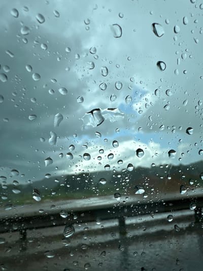 This media file captures a close-up view of raindrops on a glass surface, likely a window. The droplets vary in size, with many appearing as small spheres, while others are larger and more irregular, showing the effects of surface tension and how they distort the view behind.

The background is a blurred and muted scene, suggesting overcast or stormy weather. Patches of lighter sky with some hints of blue peek through darker, cloudy formations, creating a dramatic and somewhat somber atmosphere. To the right side of the frame, a darker, vertical element is visible, possibly a building, window frame, or vegetation, also obscured by the rain and the glass.

The light is diffused, indicating it is likely daytime, but the cloudy conditions suggest it is not bright sunlight. The overall impression is one of a rainy day, creating a sense of introspection or calm. There is no visible text in the image. The location context of Zubtsov, Russia, might evoke a sense of a rural or urban environment experiencing inclement weather. The focus is entirely on the water droplets and their interaction with light and the glass, making the abstract beauty of the rain the primary subject.