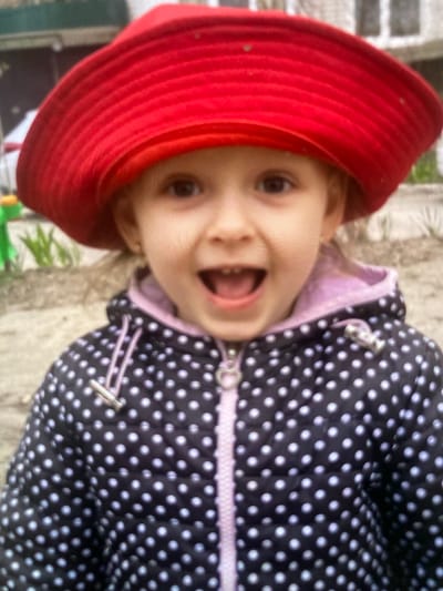 A young girl wearing a red hat and a black and white polka dot jacket is pictured outdoors, likely in a park or playground. The background suggests an urban environment with some greenery and structures. The girl is looking at the camera with a wide smile, her mouth open. The image is taken from a slightly low angle, focusing on the girl's face and upper body. The overall impression is one of a cheerful, candid moment captured in a casual setting.