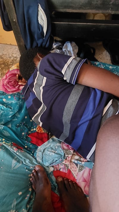 The image shows a person lying on a bed with a colorful floral-patterned comforter. The person is wearing a dark blue t-shirt with horizontal stripes of white and gray. Their head is turned away from the camera, and their left arm is bent. In the foreground, two bare feet are visible, one on the left and one on the right side of the frame. The feet are dark-skinned. To the left of the person, a pink rose-patterned fabric is visible. Behind the person, a dark blue garment with a white lining in a pocket is draped over what appears to be a dark wooden surface. Scattered on the floor around the bed are some folded clothes and a black slipper. The floor is a rough, concrete-like texture. The scene suggests a casual, indoor setting. The provided context places this scene in the city of New Nyanya, Nigeria.