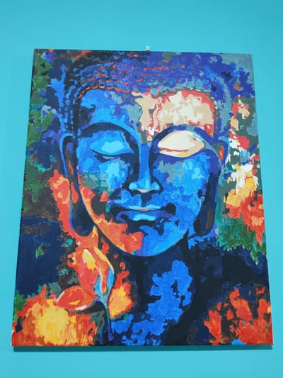 The image shows a painting of a Buddha's face rendered in a vibrant, abstract style. The face is primarily depicted in shades of blue, with accents of red, orange, yellow, and cream. The eyes are closed, and the expression is serene. Surrounding the face are abstract shapes and colors that suggest foliage and flowers, rendered in blues, greens, reds, and yellows. The painting is hung on a turquoise wall. There are no people or animals visible in the image. The location is not discernible from the visual information provided.