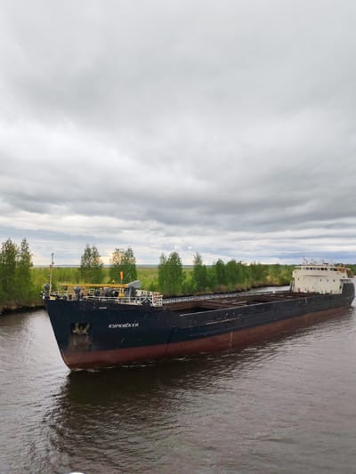 A barge, identified with the name "КУРКИЁККИ" painted in white on its bow, is moving through a waterway in what appears to be Ozernoe Ust'e, Russia, given the style of lettering on the ship. The vessel is long, black and red, and appears to be empty. The water is dark and choppy. The barge is navigating past a bank lined with green trees and foliage. Overhead, the sky is overcast with gray clouds.
