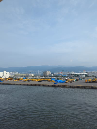 The image depicts a waterfront industrial area in Kobe, Japan, likely within the port district. The scene is dominated by a large paved lot filled with rows of yellow construction equipment, including excavators and what appear to be truck beds. Several large blue tarpaulins are draped over piles of materials. In the background, buildings of various sizes and architectural styles are visible, including what looks like a large factory or warehouse with a white arched structure and several multi-story buildings. Further in the distance, a mountain range forms the backdrop, partially obscured by haze. Two large ferries, one with a red and white upper section, are docked in the harbor. The water in the foreground is dark and choppy, indicating a moderate level of activity or wind. The sky is a pale blue with scattered clouds, suggesting daytime. There are no people clearly visible in the frame. The overall impression is one of a bustling port and industrial zone dedicated to construction and shipping.
