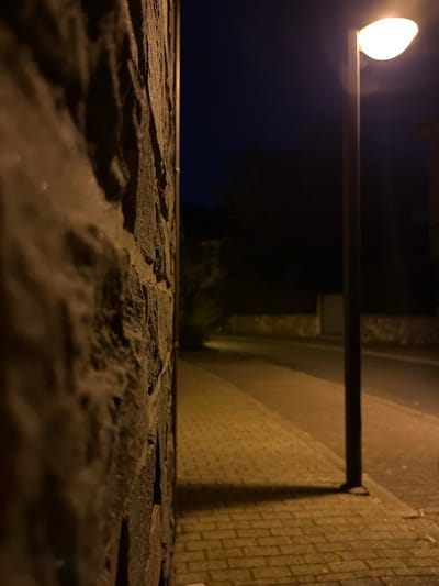 The image depicts a night scene on a street in Saint-Vith, Belgium. The foreground is dominated by a rough-textured stone wall on the left, with its surface heavily shadowed and out of focus, showing variations in its stony texture. To the right, a paved sidewalk runs parallel to a street, illuminated by a single street lamp. The street lamp, with a white, oval-shaped light, is positioned on the right side of the image, casting a distinct shadow onto the sidewalk. The sidewalk itself is paved with rectangular bricks that are lit by the lamp, creating a warm, orange glow. The street beyond the sidewalk is dark and appears to be empty. In the background, silhouetted trees and buildings are faintly visible against the dark night sky, suggesting a residential or commercial area. The overall atmosphere is quiet and deserted.