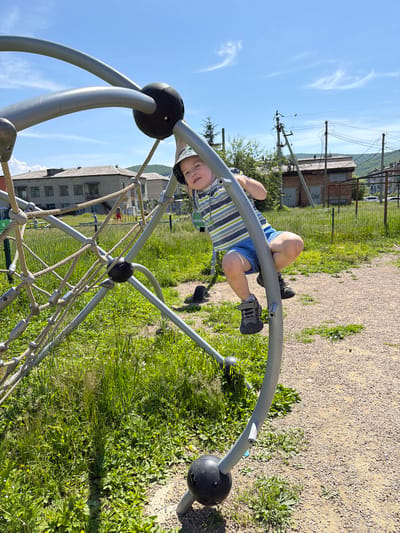 A young boy wearing a striped t-shirt, blue shorts, a bucket hat, and dark sneakers climbs on a geodesic dome jungle gym. The jungle gym is made of grey metal bars and ropes, with black spherical connectors. The boy is positioned on the right side of the jungle gym, with his legs bent and his hands gripping the metal bars. His face is turned towards the camera, and he appears to be smiling. The background features a grassy area with scattered yellow flowers, a gravel path, and a chain-link fence. In the distance, several buildings, including a two-story apartment building, and utility poles are visible. The sky is clear blue with a few wispy white clouds. This appears to be a public playground in the rural settlement of Kul'dur, Russia.