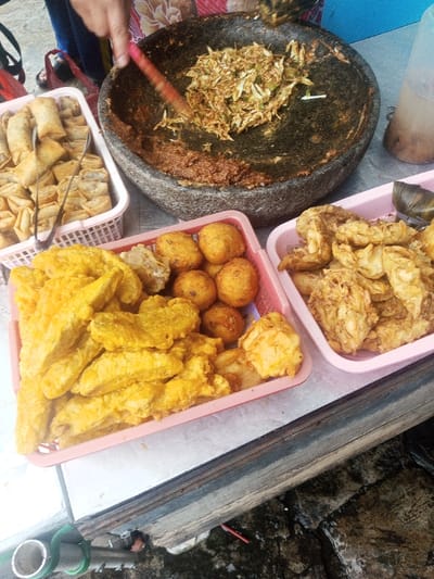A street food vendor prepares gado-gado in a stone mortar. Fried snacks are arranged in pink plastic baskets: spring rolls, tofu, fried bananas, fried vegetable balls, and other unidentified fried foods. Tongs are resting in the spring roll basket. A tall jar containing liquid is visible on the right side of the image.