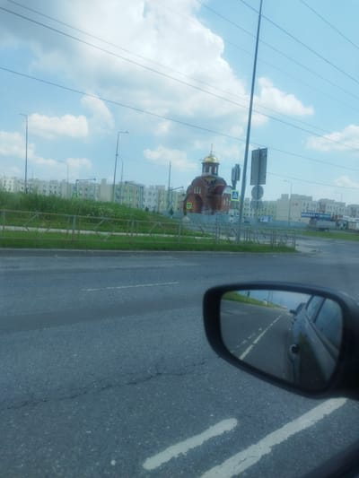 The image, taken from a vehicle, shows a street scene in Stroykeramika, Russia. A modern-style church with a golden dome is visible on the left, behind a metal fence and a grassy area. To the right of the church are multi-story residential buildings. The sky is blue with scattered clouds. Power lines are prominent across the top of the frame. In the foreground, the asphalt road has white lane markings. The side mirror of the vehicle reflects the road and other vehicles behind it.