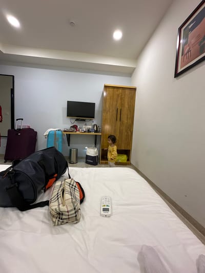 The scene is an indoor hotel room located in Vadodara, India. The perspective is low, looking towards a wall furnished with a desk, television, and wardrobe.
In the foreground, a large white bed occupies the lower half of the image. Resting on the white duvet are a dark blue duffel bag with orange accents, a beige plaid toiletry bag, and a white remote control. A white towel is partially visible on the far right edge of the bed.
In the midground, a light-skinned child, approximately 1-2 years old, with dark curly hair, wearing a yellow long-sleeved shirt and light-colored pants, stands near a tall wooden wardrobe on the right. The child is facing and looking into an open lower compartment of the wardrobe, which contains a folded yellow object. To the left of the wardrobe, a black flat-screen television is mounted above a wooden floating shelf or desk. On the shelf are multiple plastic water bottles, a black electric kettle, and other small items. Below the desk, a silver cylindrical trash can and two shopping bags, one white with "JADEUE" text and one yellow, are on the floor. Further left, a light blue wheeled suitcase is positioned against a light blue wall, next to a larger maroon wheeled suitcase.
The walls are predominantly light blue on the left and white on the right. A framed abstract artwork featuring pink and blue shapes is hung on the white wall. The white ceiling has multiple recessed circular spotlights and a perimeter light strip. A dark doorframe is partially visible on the far left. The floor appears to be a light-colored tile or wood laminate.