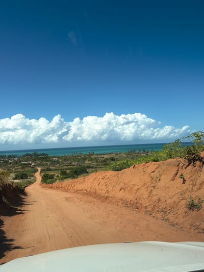 A dirt road curves through a sandy, arid landscape under a bright blue sky dotted with large, fluffy white clouds. The road is bordered by a steep, orange-red dirt embankment on the right and sparse vegetation on the left. In the distance, the turquoise ocean meets the horizon, fringed by a line of green trees and some scattered buildings. The scene appears to be a rural area in Muxara, Mozambique, likely a coastal region given the ocean view. The perspective suggests the photo was taken from the front of a vehicle.