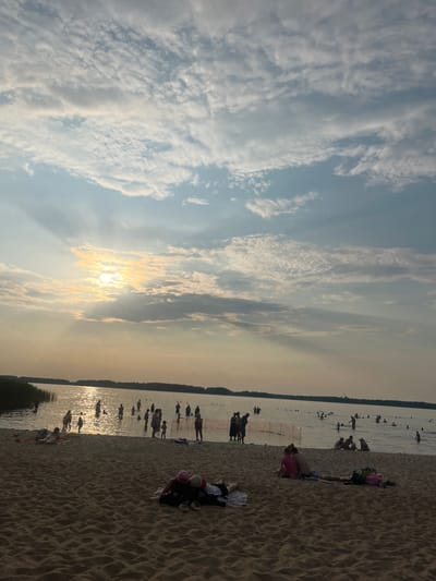 The image captures a scene at a sandy beach with people enjoying the water and the shoreline. The sky is filled with a mix of bright clouds and streaks of sunlight, suggesting it is either sunrise or sunset. The sun's rays are prominently visible, shining through the clouds and reflecting on the water.
Several individuals are visible in the water, some appearing to be swimming or wading. On the sandy beach, other people are seen relaxing, sitting, or lying down. Some are engaged in conversation or simply enjoying the beach atmosphere. The sand is textured with footprints and ripples, indicative of a well-used beach.
The overall mood appears to be peaceful and recreational, with people spending a pleasant day outdoors. The water is calm, and the distant shoreline is lined with trees, providing a natural backdrop. The specific location is identified as Загародскі сельскі Савет, Belarus. There is no visible text in the image.