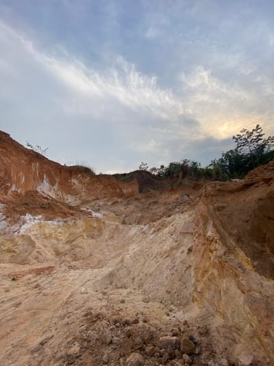 An expansive open-air excavation site is depicted under a partly cloudy sky in Asaga Amangwu, Nigeria. The terrain consists of exposed, stratified earth, exhibiting distinct reddish-brown, orange, and lighter beige-white layers. The landscape is characterized by steep, irregular slopes and an uneven, excavated floor composed of loose soil and rock fragments. Minimal vegetation, comprising small trees and shrubs, is visible along the upper edges of the excavation perimeter. The sky presents a mixture of grey and white clouds, with a discernible yellowish light source in the upper right quadrant. No individuals, human activity, or man-made objects are present within the scene.