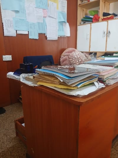 A messy desk displays scattered papers, a blue cloth item, and a black object. A thick stack of files and documents leans against the desk's edge, next to a plush pink cushion. A white power outlet is visible near the desk's corner. A wooden shelf with drawers stands nearby. The desk itself is made of wood and has a slight curve on one side. A glimpse of the floor is visible under the desk.