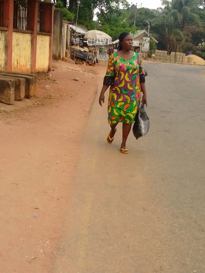 This image depicts an outdoor street scene in Umuoji, Nigeria, under overcast conditions. In the foreground, a dark-skinned adult female walks from left to right on an asphalt road, facing slightly right of the camera's perspective. She wears a vibrant, short-sleeved dress with a green, purple, yellow, and orange floral or leaf pattern, black and white polka dot cuffs, a dark headscarf, and yellow flip-flops. Her left hand holds a dark-colored plastic bag.
To the immediate left of the asphalt road is a reddish-brown dirt shoulder. Along this shoulder, in the midground, a building with a yellow base and reddish-brown upper section, featuring a metal-barred window, is visible. Three large, rectangular concrete blocks are stacked on the dirt in front of this building. Further down the road on the dirt shoulder, several market stalls or small shops are present, one with a white canopy and a "POS" sign. Another individual is faintly visible near these stalls. In the background, stacks of red bricks or concrete blocks are piled on the right side of the road, and abundant green trees and vegetation line the horizon. Utility poles and power lines are visible overhead. The overall environment suggests a bustling, semi-urban or rural commercial area.