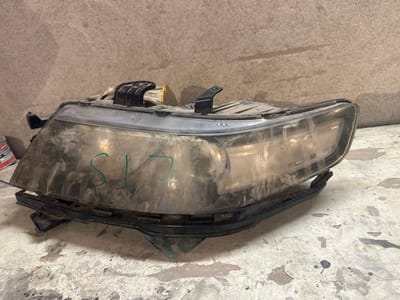 A dusty, used car headlight assembly sits on a speckled concrete surface in what appears to be a workshop or garage. The headlight is made of clear plastic and black plastic components, with visible scuffs, dirt, and what appears to be green marker writing on the lens that reads "SL 17". The background is a textured, light brown concrete wall. No people are visible in the image, and there is no information to indicate a specific location within International, Kyrgyzstan.
