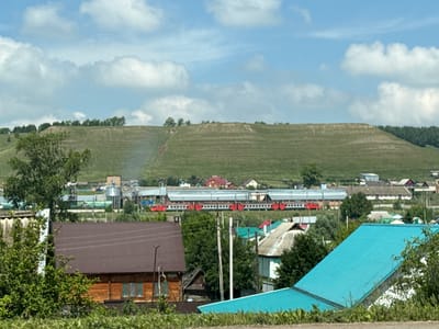 The image depicts a rural landscape with a village in the foreground and a large, terraced hill in the background. A red and white train is visible on tracks running horizontally across the mid-ground, passing in front of several industrial-looking buildings. In the foreground, there are several houses with brown and bright blue roofs, surrounded by greenery. The sky is blue with scattered white clouds. The scene appears to be set in a somewhat developed, but predominantly natural, environment. The location is identified as the city of Kandry, Russia. There are no people or animals visible in the image.