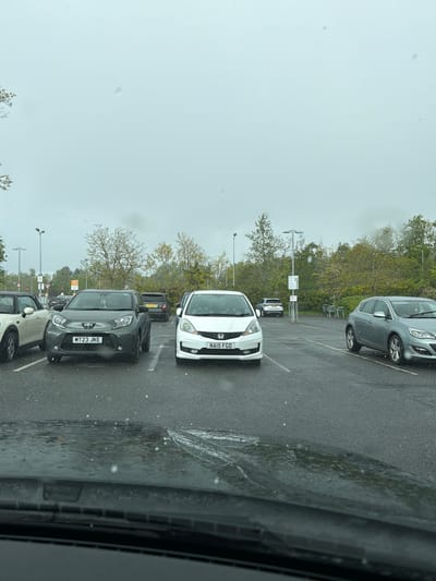 The image shows a parking lot in Pity Me, United Kingdom on a rainy day. Multiple cars are parked in rows. A white Honda with the license plate "NA15 FGO" is prominently visible. Behind it is a dark car and in front of the Honda is a grey Toyota with the license plate "MT23 JKE". Other vehicles are also parked nearby. The trees in the background suggest it is near a wooded area. The image was taken from inside a car.