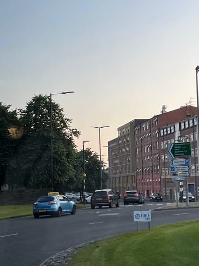 The image shows a street scene in the city of Derry/Londonderry, United Kingdom, with several cars driving on a road. In the foreground, a blue Renault Megane taxi is visible. In the middle ground, several other vehicles, including SUVs, are driving. In the background, there are buildings and trees. A green directional sign points towards "Derry" (Belfast A6), "Larne", "Coleraine A2", and "Waterside". Another sign points to "Foyle Road" and "Multi-Storey Car Park". A smaller sign on the grass reads "FOYLE PORT". The sky is a pale, hazy orange, suggesting it is either sunrise or sunset.