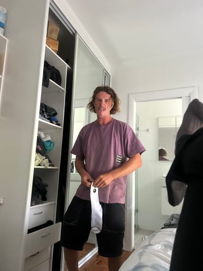 A young man with long hair is sticking out his tongue, posing in front of a mirrored closet. He wears a mauve t-shirt and black shorts, holding a white sock with a black detail in his hands. The closet behind him has sliding mirrored doors and is filled with clothes and storage boxes. A bathroom can be seen in the mirror's reflection. A sock and part of a bed obscure the top of the frame.