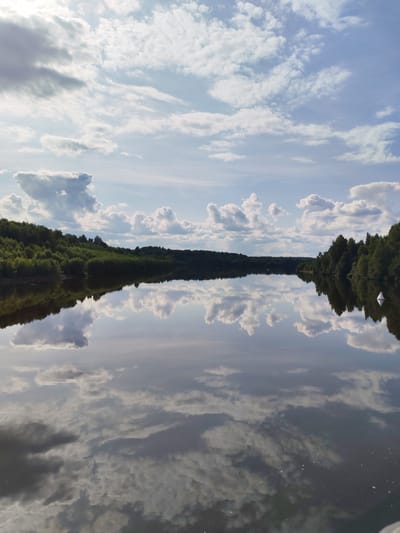 A wide, calm river reflects a bright blue sky filled with fluffy white clouds and dappled sunlight. Lush green trees line the distant banks of the river, creating a dense forest that extends as far as the eye can see. The reflection in the water is nearly perfect, mirroring the sky and the trees with remarkable clarity. A white buoy is visible in the distance on the right side of the river. The scene evokes a sense of peacefulness and natural beauty, characteristic of a serene landscape in Srednii Rubezh, Russia.